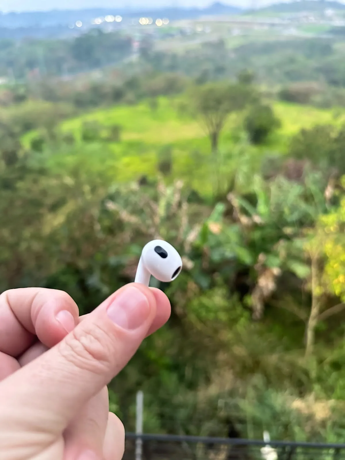 Fone de ouvido branco segurado entre os dedos, com vegetação e um vale ao fundo