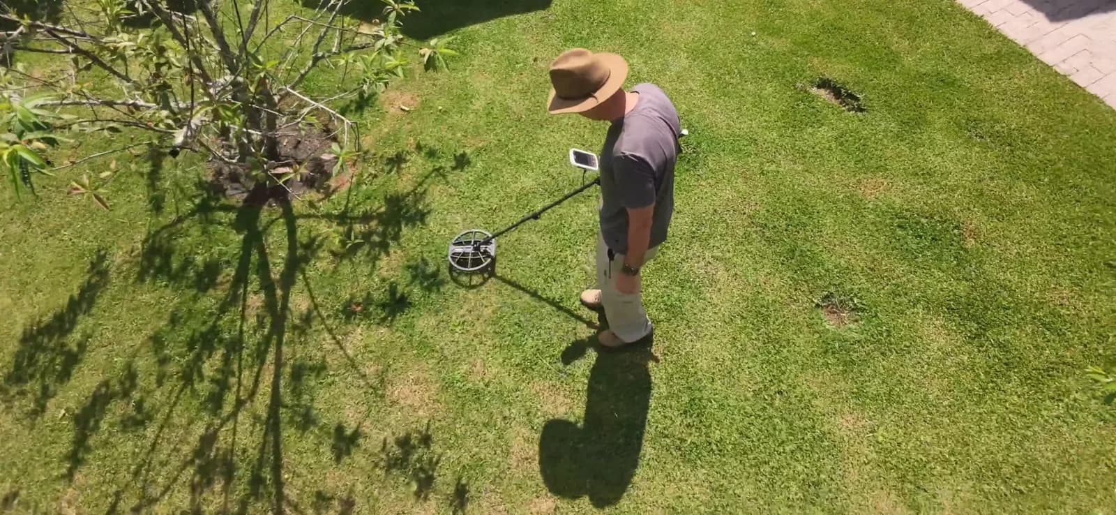 Detectorista de chapéu passando o detector de metais sobre um gramado baixo, visto de cima